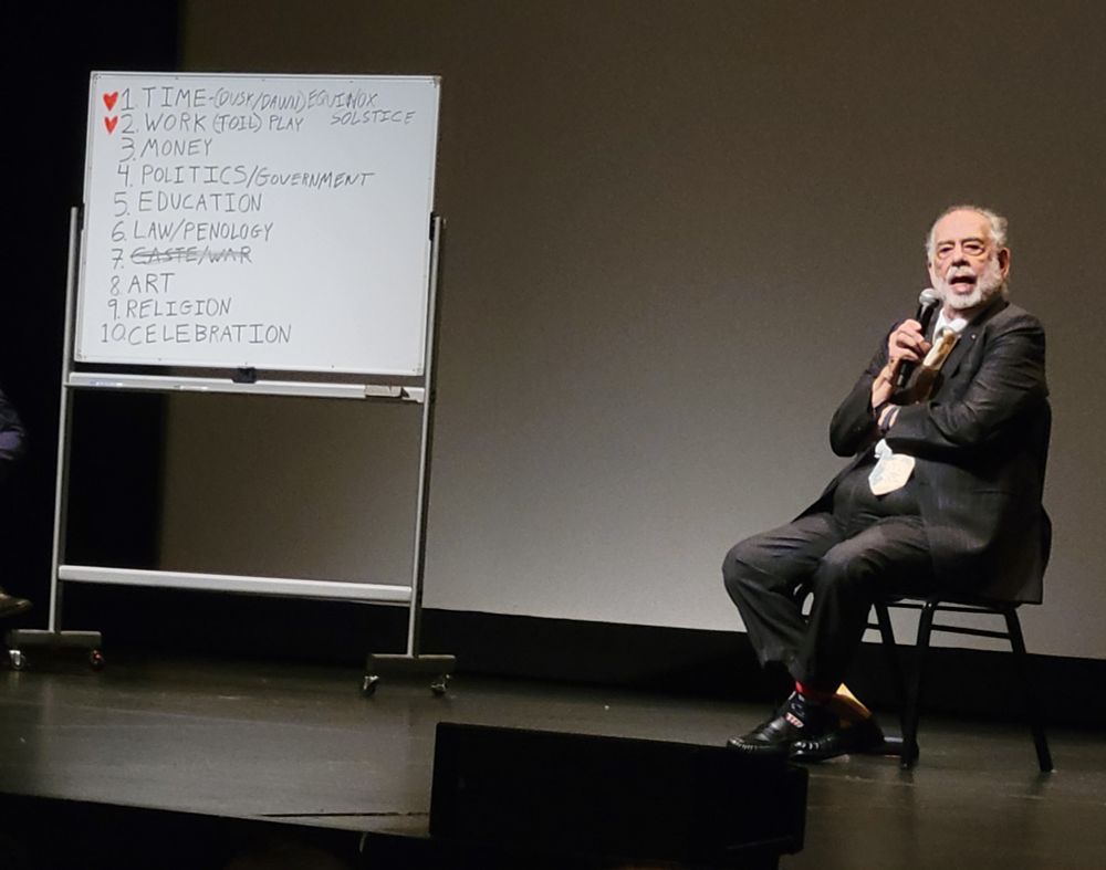 Photo of seated Francis Ford Coppola speaking into a handheld microphone. He wears a charcoal-colored suit, white shirt, black loafers, mismatched colorful socks, and a yellow silk tie. At left is a portable whiteboard with a list of 10 items: ❤️ Time (Dusk/Dawn) Equinox/Solstice, ❤️ Work, Money, Politics/Government, Education, Law/Penology, (crossed-out) Caste/War, Art, Religion, and Celebration.