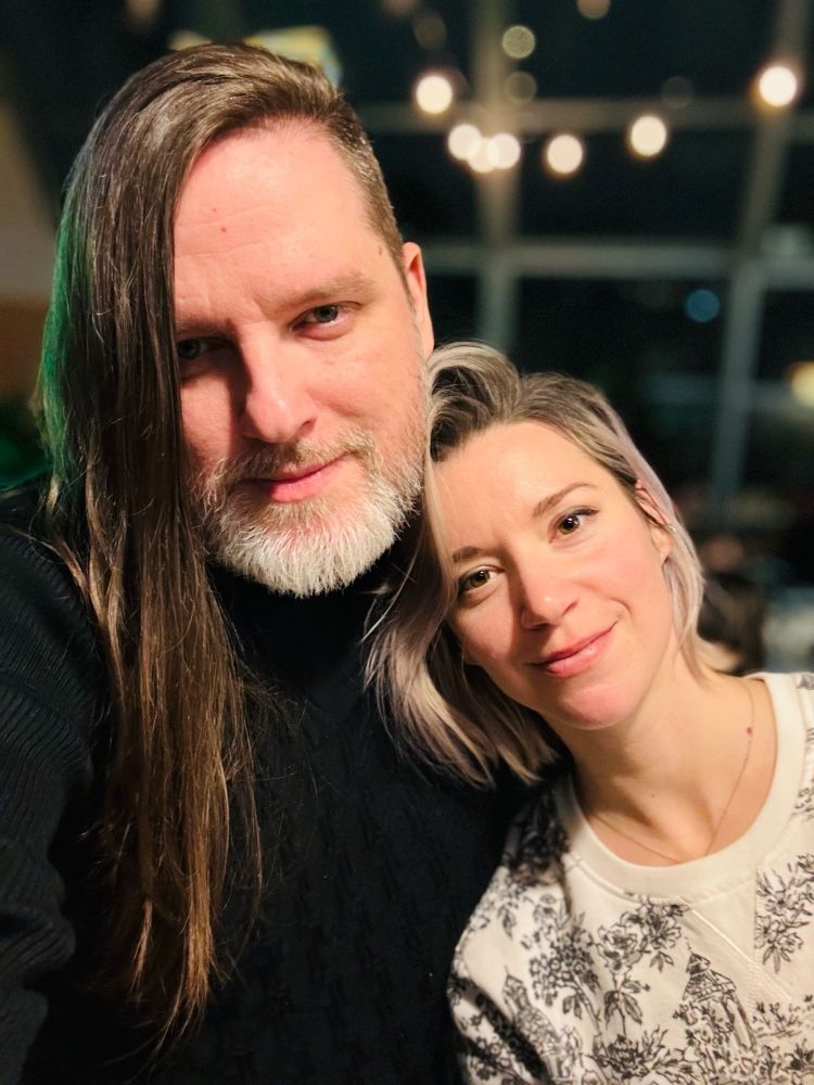 Laurin and Robert, aka: Sphynxian, leaning together and smiling at the camera, in a dimly lit restaurant. 