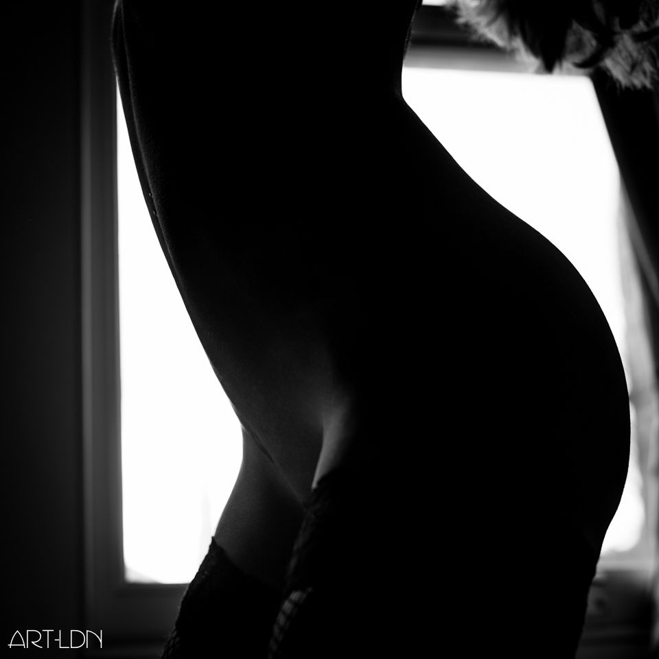 We see a black and white image of a fitness model, composed to capture her from mid thigh to just below her breasts. She is naked apart from stockings which are pulled high up her toned thighs. She's framed kneeling on a bed in front of a window, almost entirely silhouetted apart from a little light that dances across the front of her thighs and lower stomach.