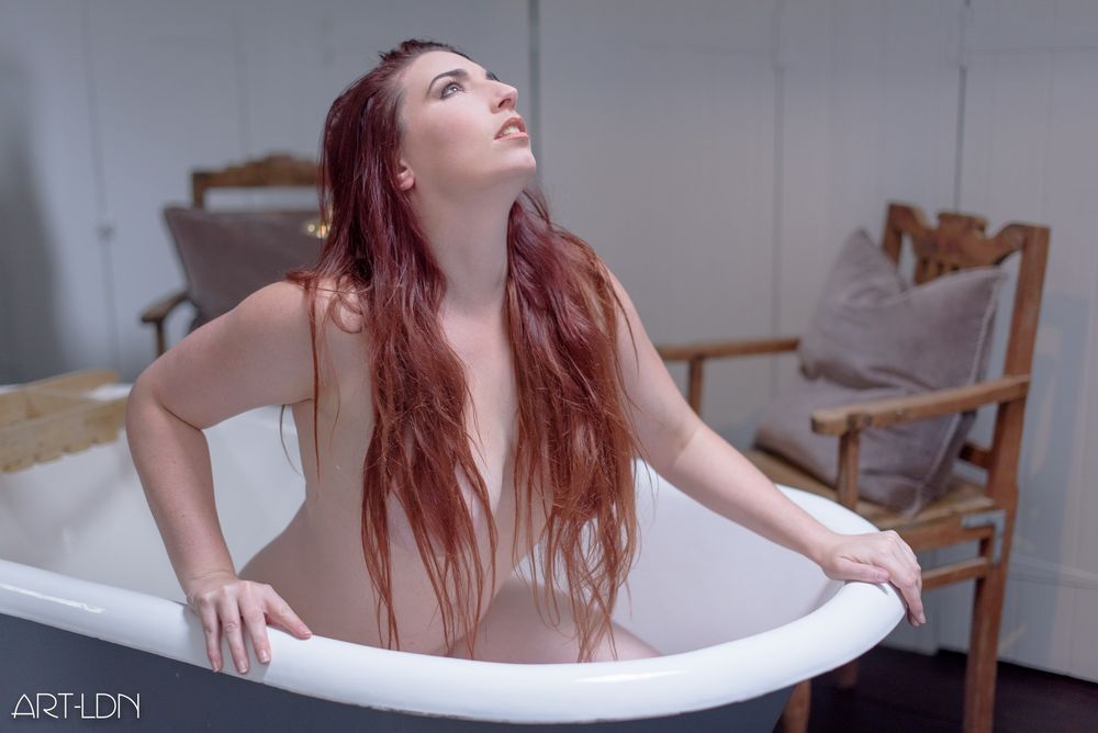 We see a female redhead model in a bathtub. She's nude but her hair covers her breasts as she crouches, poised to rise from the tub for her day. She tilts her head to the light which comes from a skylight above her head.