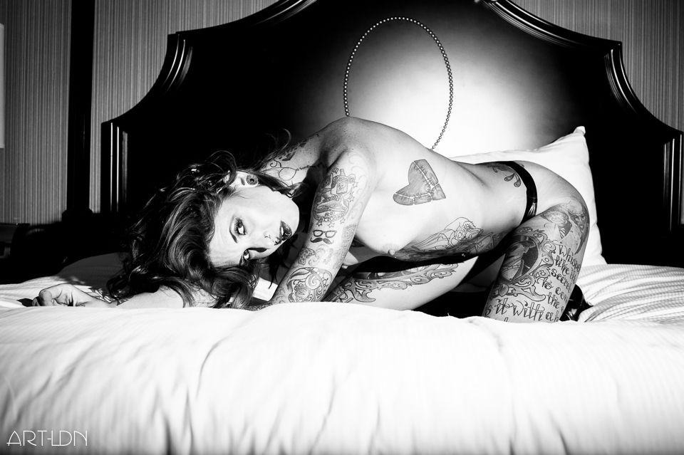 A noir inspired boudoir art nude image, with a redhead kneeling on a bed in a luxurious hotel. She leans forward on her elbows, looking at us with a challenging and intense gaze. She's heavily tattooed and wears a small laced latex thong.