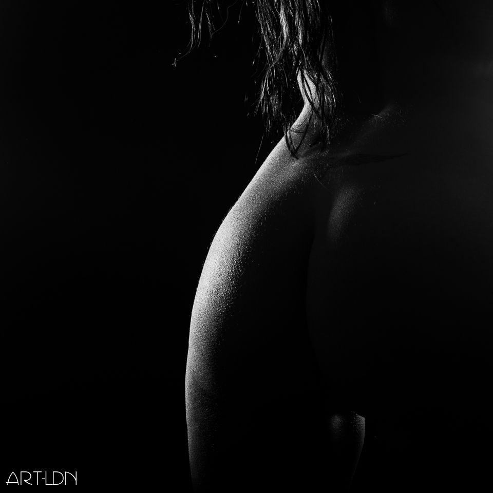 A black and white image taken in a studio of a nude fitness model. We see her from behind, close up to show the top of her thighs to just above the small of her back. A little hair cascades down into shot. The curve of her hip is lit up but most of her body is in shadow, other than a little light on her inner thigh, leaving much up to the viewer's imagination.