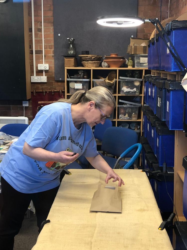Woman carefully adjusting something inside a brown paper bag on the hessian table. Above her head is an illuminated ring light with a phone camera in the centre and there is a tiny remote control in her hand.