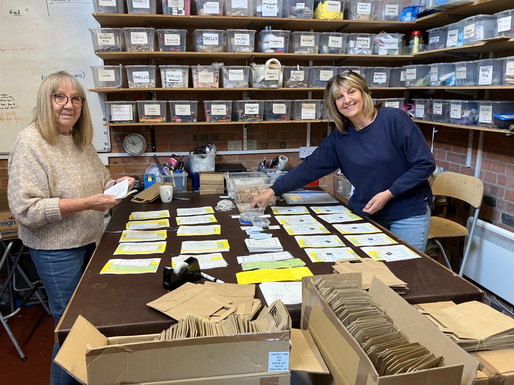 Two ladies smile at the camera. They are either side of a large table where many identical stacks of coloured card, googly eyes, etc are being laid out. The walls of the workshop are lined with shelves of boxes of craft resources. In the foreground, cardboard boxes contain stacks of filled paper bags.
