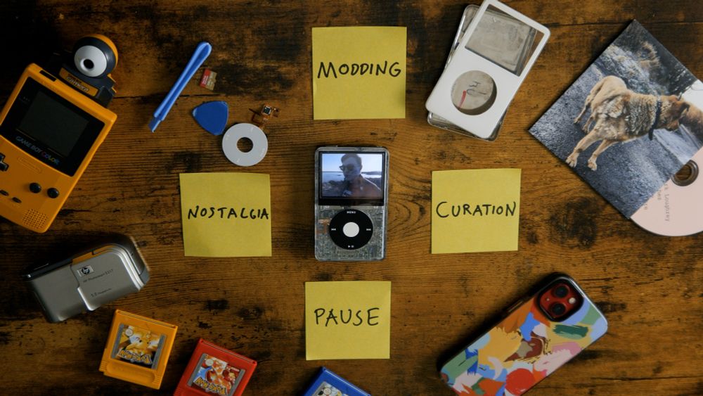 A model 5 ipod with a transparent shell on a table surrounded by post it notes that read PAUSE, CURATION, MODDING and NOSTALGIA. Also on the table are a CD, a smartphone, the shell and some innards of the gutted iPod, a Game Boy with Pokémon games and a Game Boy Camera, and an old, digital camera.