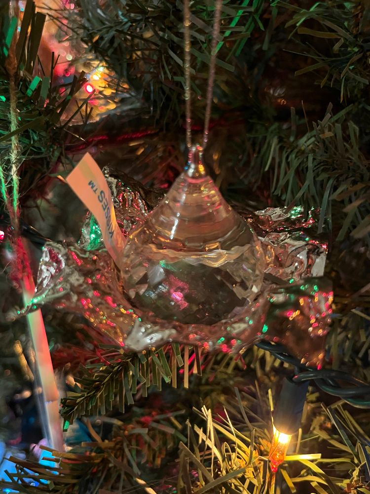 A picture of a Christmas ornament that’s a Hersheys’s Kiss made out of glass with the silver wrapper and tag