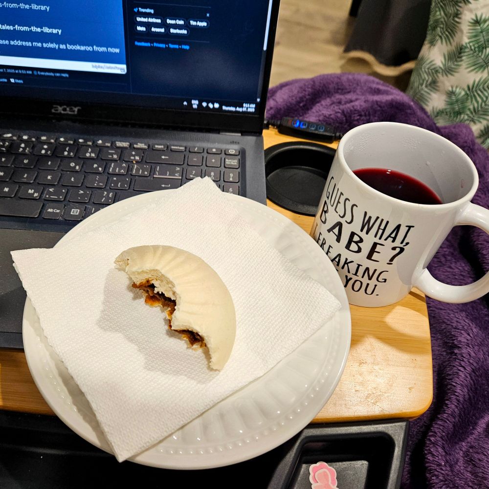 A photo of my swivel desk, with part of my laptop visible. Next to it is a half-eaten pork bun and a mug that says, "Guess what, babe? I freaking adore you."