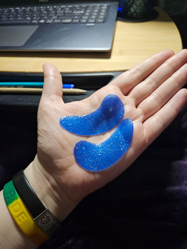 My hand, where a pair of bright blue under-eye masks are displayed on my open palm. They are absolutely resplendent with embedded multicoloured glitter.