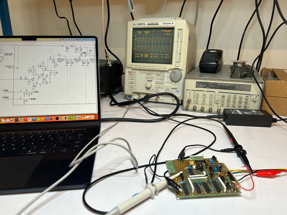 Workbench with laptop showing a schematic, an oscilloscope showing a square waveform and a sine wave, and a PCB with probes and wires. 