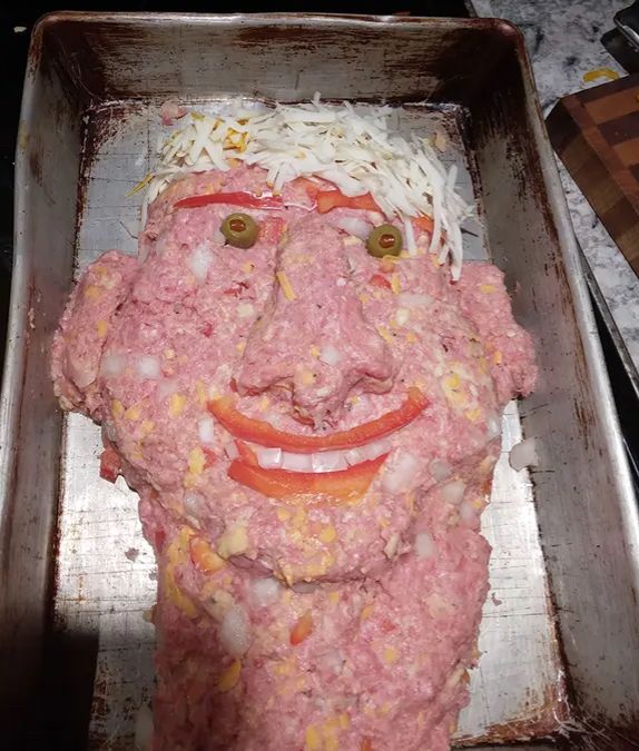 A meatloaf shaped like a head with shredded cheese, hair, red pepper, lips, and eyebrows, pimento, olive eyes and onion teeth
