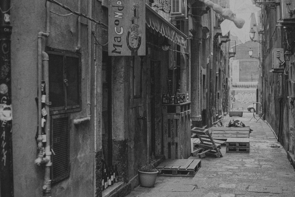 Schwarz-Weiß-Fotografie einer schmalen Gasse in Catania, Sizilien. Im Vordergrund sind improvisierte Sitzgelegenheiten aus Europaletten und verlassene Flaschen zu sehen. Die alten, verwitterten Häuserwände säumen beide Seiten der gepflasterten Gasse. Ein Schild mit 'Macondo' hängt an der linken Wand. Am Ende der Gasse öffnet sich der Blick auf einen helleren Platz. Die Nachmittagssonne wirft weiche Schatten und betont die rauen Texturen der historischen Architektur. Die Szene vermittelt eine ruhige After-Party-Atmosphäre in einer charakteristischen italienischen Altstadtgasse.