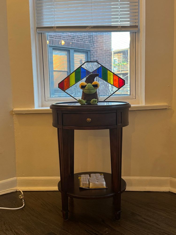 a frog wizard plush by elmrealm sitting on a small circular wooden side table in front of a window with a piece of rainbow stained glass art behind it