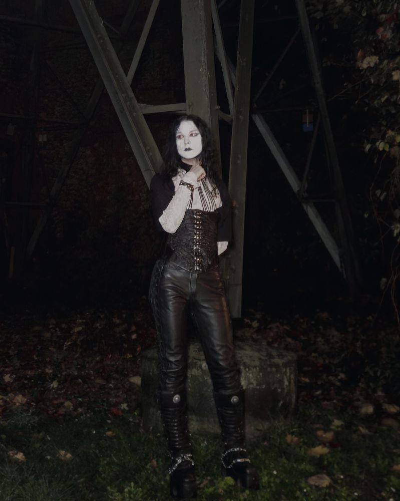 I am standing with my hand on my choker. I am wearing red contacts. I am wearing a white lace shirt with an open black button-up shirt. I am wearing a corset with a choker. My pants are black leather pants 