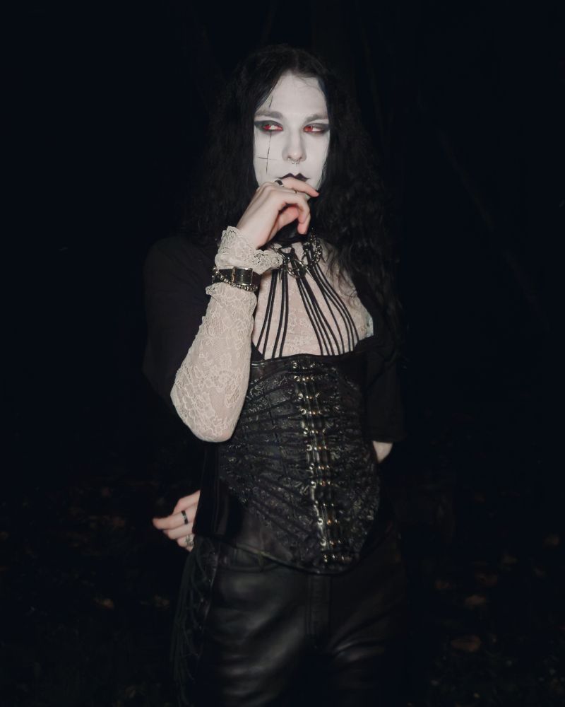 I am standing with my hand in front of my mouth. I am wearing red contacts. I am wearing a white lace shirt with an open black button-up shirt. I am wearing a corset with a choker. My pants are black leather pants 