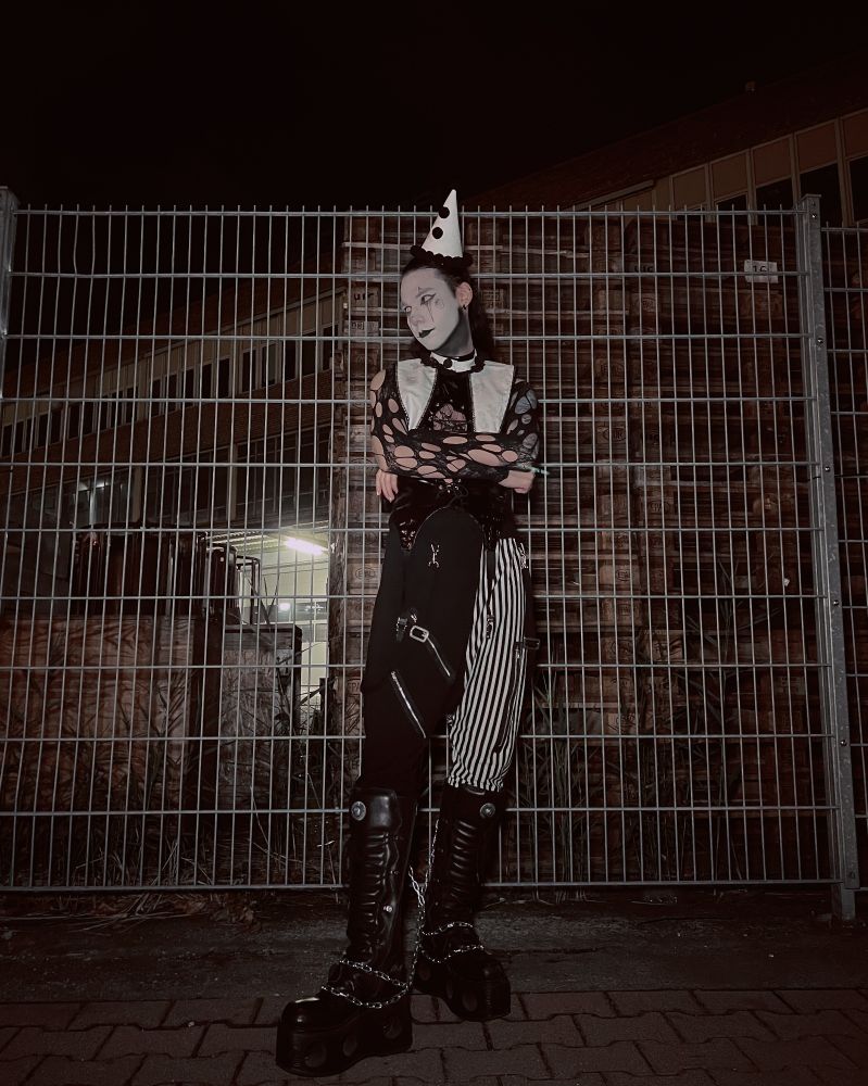 Picture of me in a goth clown outfit in front of a metal fence. I am wearing a black and white party hat and collar. I am wearing a black vinyl top and fishnets over my arms. My pants are black on one leg and vertically striped black and white on the other. I am wearing platform boots with chains on them 
