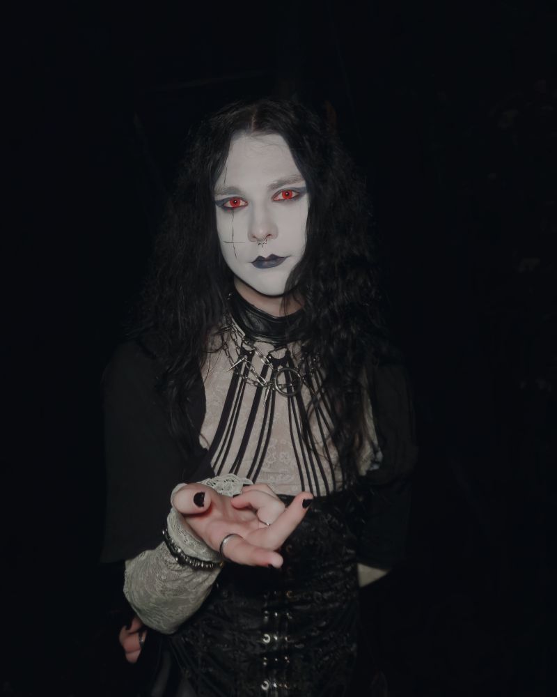 I am standing with my hand extended forward. I am wearing red contacts. I am wearing a white lace shirt with an open black button-up shirt. I am wearing a corset with a choker. My pants are black leather pants 