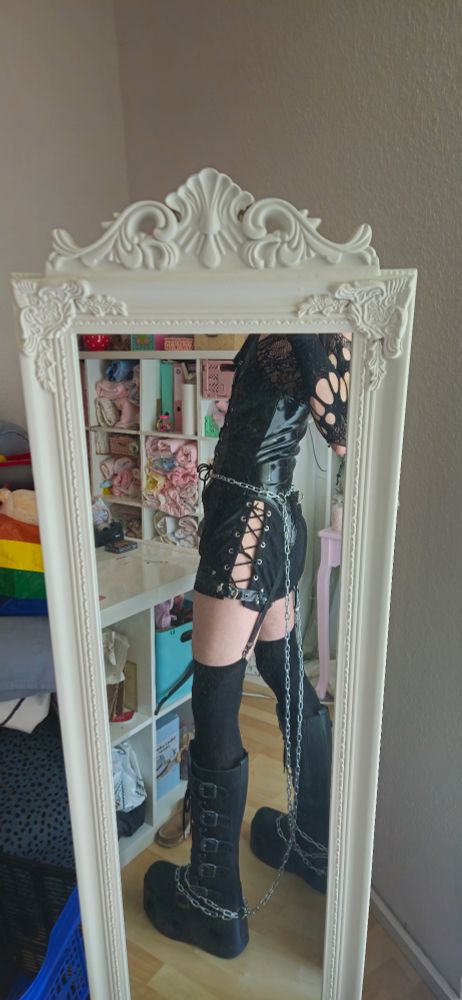 Mirror selfie of me wearing shorts with open sides a vinyl top that is strapped to overknee socks and I am wearing platform boots
