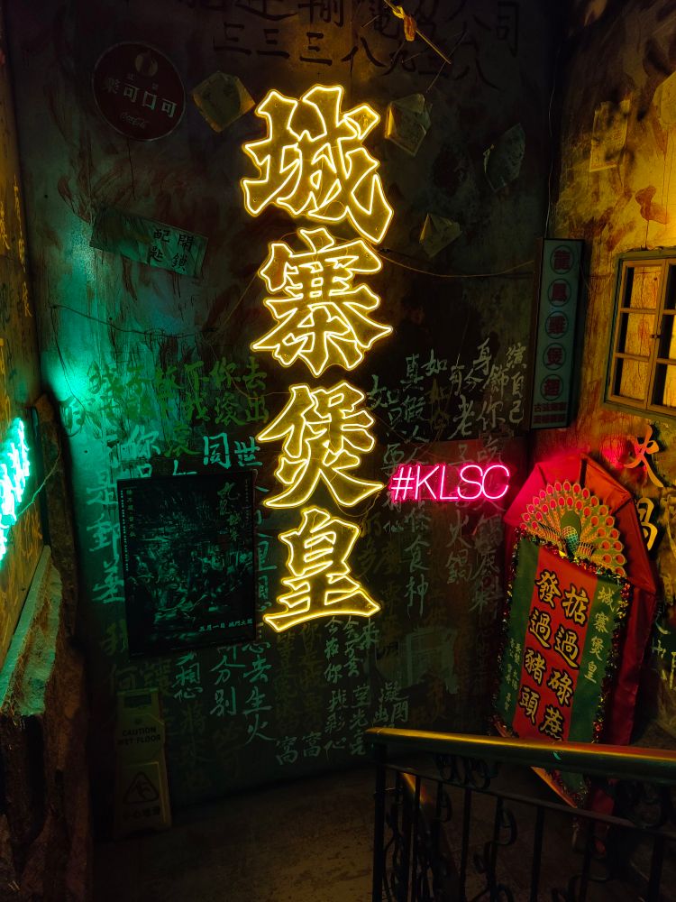 城寨煲皇の階段踊り場。黄色いネオンの店名が輝く。ネオンの横には九龍城寨之圍城ポスターが飾られている。
