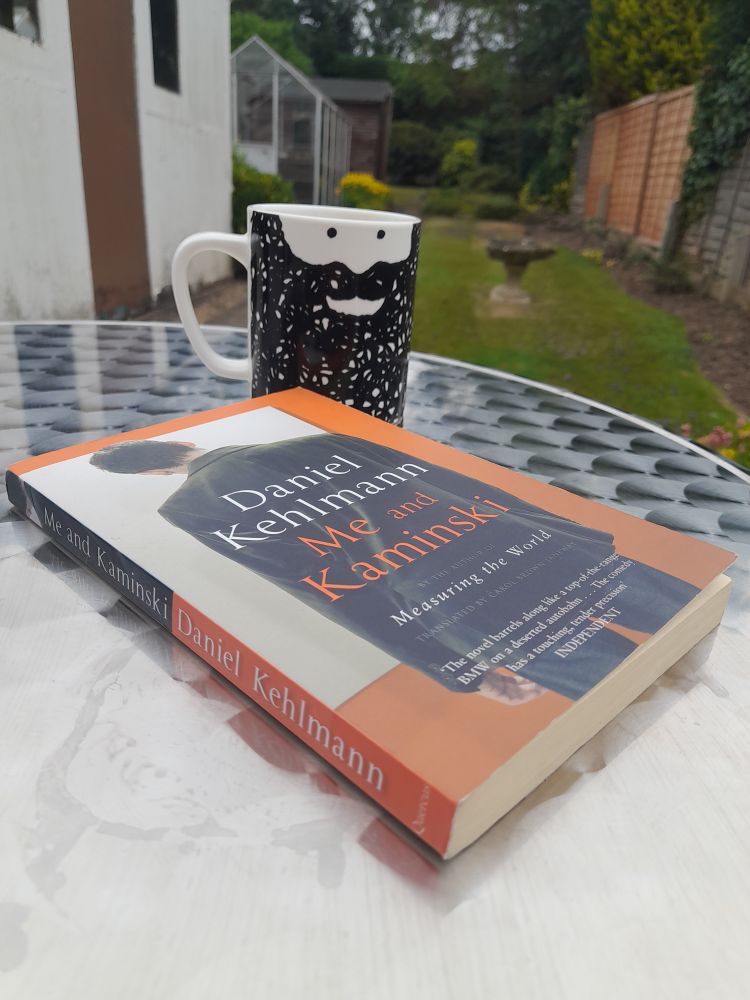 Paperback copy of 'Me and Kaminski' by Daniel Kehlmann, on a patio table next to a (beardy) mug. Tea is mango & lychee green tea fwiw. Garden pictured in the background has seen sunnier days, and will again.