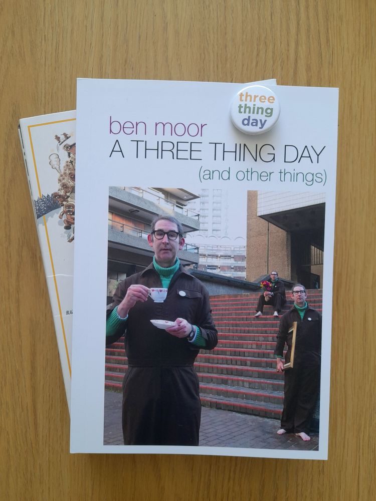 A paperback copy of Ben Moor's new book, 'A Three Thing Day (and other things)', and a 'Three Thing Day' pin badge, pictured on a coffee table. Underneath the book is a BluRay copy of Hundreds of Beavers, because why not. The cover of 'ATTD' is white with a large photo showing three versions of the author in different poses, on/near steps leading up to a Brutalist building.