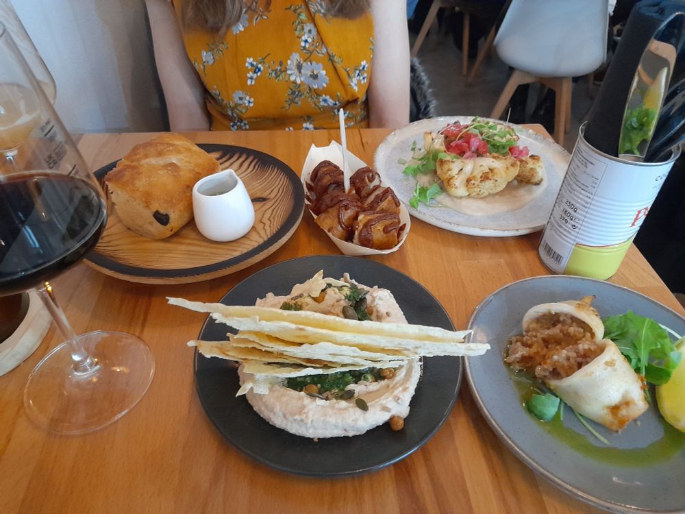 On top of a bar table, there is a glass of dark beer and various foodstuffs served on small plates, including currywurst, cauliflower, stuffed squid, focaccia and humous.