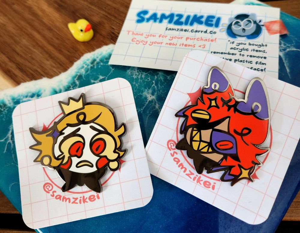 image is a photograph of two enamel pins featuring the the faces of the characters Lucifer Morningstar and Alastor from the series Hazbin Hotel. A small business card sits on the wooden surface just above them next to an exceedingly tiny rubber duck. #hazbinhotel