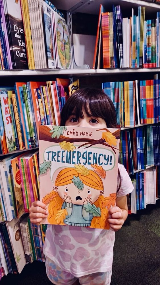 A little girl standing in a bookshop holding up a copy of Treemergency! A picture book written and illustrated by Emily House