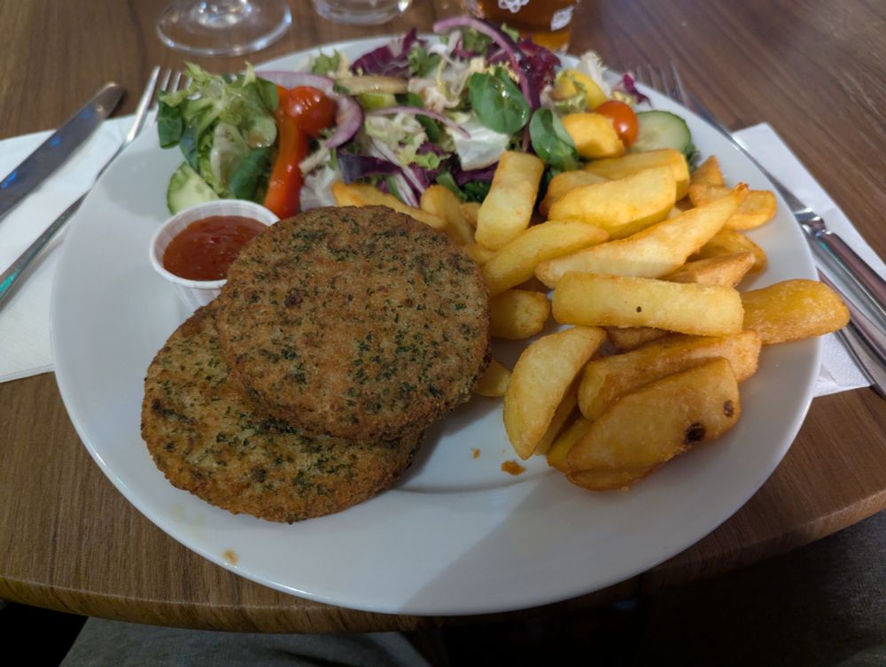 A plate of two salmon and dill fishcakes, chips, and side salad served alongside cutlery and glasses on a wooden dining table. 