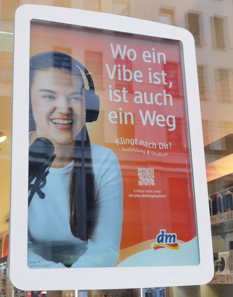 Foto von einem DM Plakat im Schaufenster. Das Plakat ist weiß gerahmt, auf weißer Schrift steht vor orangem Hintergrund geschrieben: "Wo ein Vibe ist, ist auch ein Weg. Klingt nach dir? Ausbildung & Studium". Darunter ist ein QR Code und das dm Logo. Auf der linken Plakat Hälfte ist eine junge Frau mit weißem Pullover, langen dunklen Haaren, großen Kopfhörer und Mikrofon abgebildet, sie lacht und ihr Blick geht seitlich nach vorne. 