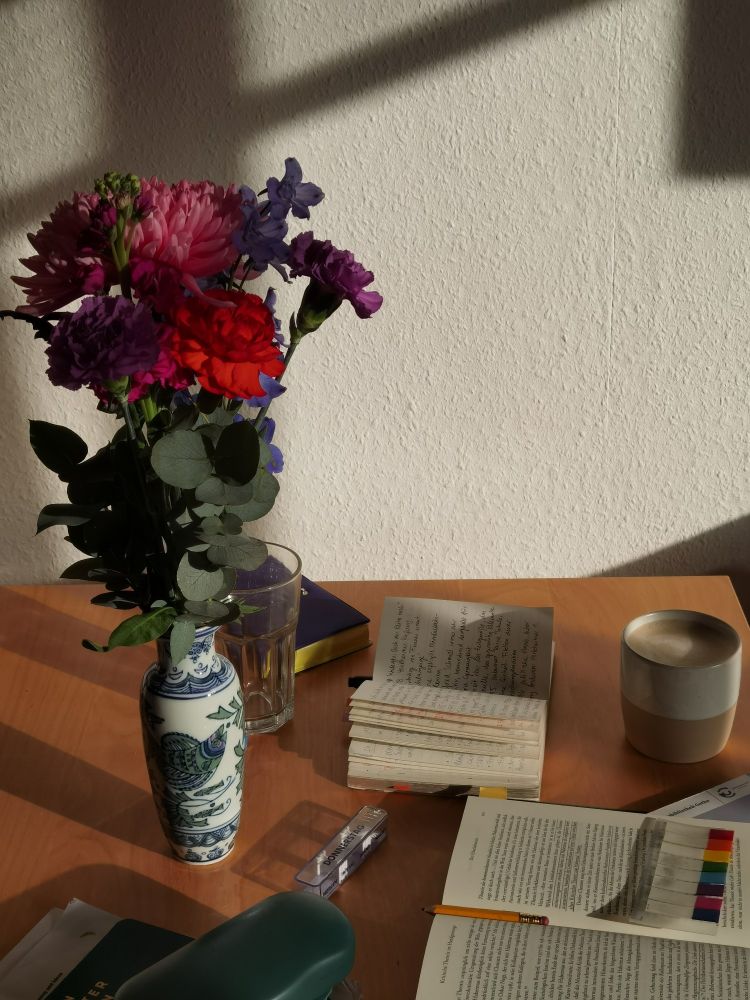Foto im Hochformat, die untere Bildhälfte zeigt einen Holztisch, auf dem ein Buch, Notizbuch, Kaffeetasse, Brillenetui, Medikamentenetui, Wasserglas und eine schmale Blimenvase versammelt sind. Die obere Bildhälfte zeigt eine weiße Wand, auf die die Sonne scheint und wenige schwarze Schattenlinien den Fensterrahmen projizieren. In die obere Bildhälfte ragt aus der Vase ein Blumenstrauß 5-8 Blüten in rot, lila und rosa Farbtönen.