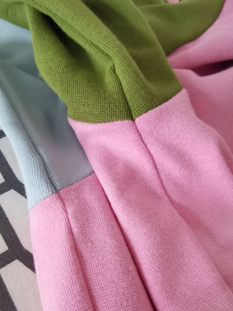 Nahaufnahme von den Achselnähten eines t-shirts. Der Körperteil ist rosa, der eine ärmel hellblau und der andere ärmel grün. An der Achselnaht treffen die Farben in einer exakt geraden Linie aufeinander. Der Stoff ist ein etwas dickerer Baumwolljersey