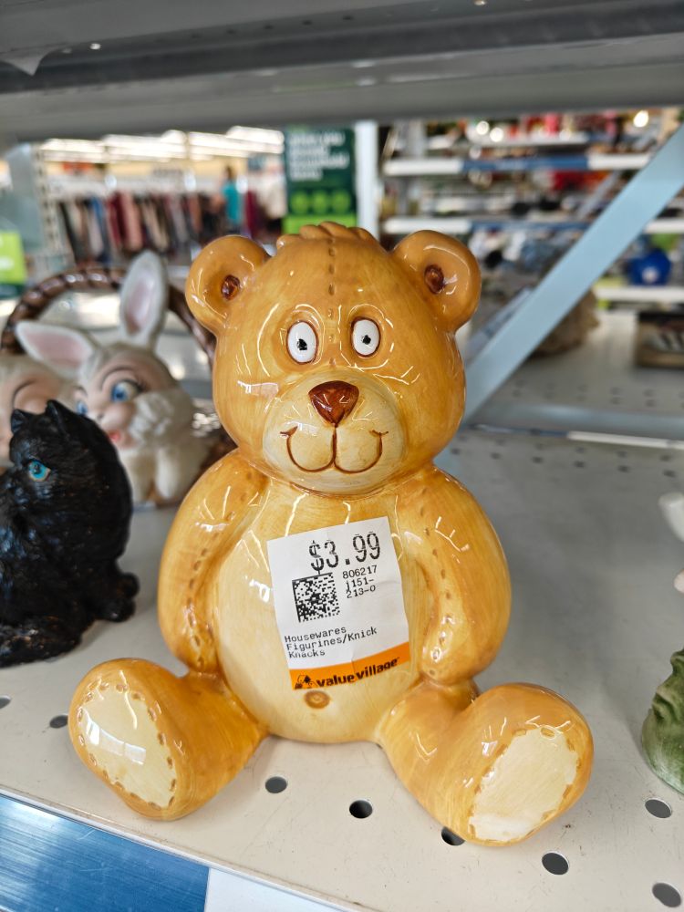 Pic of small, glazed, clay figurine of a light brown bear, on a shelf at a thrift shop.  The bear is sitting upright with it's back legs wide out front,  and has a bit of a blank expression except the eyes are completely wide open, staring at you.  Its snout design makes it look like it's smiling.  A sticker on the front of it shows a price of $3.99 (USD).