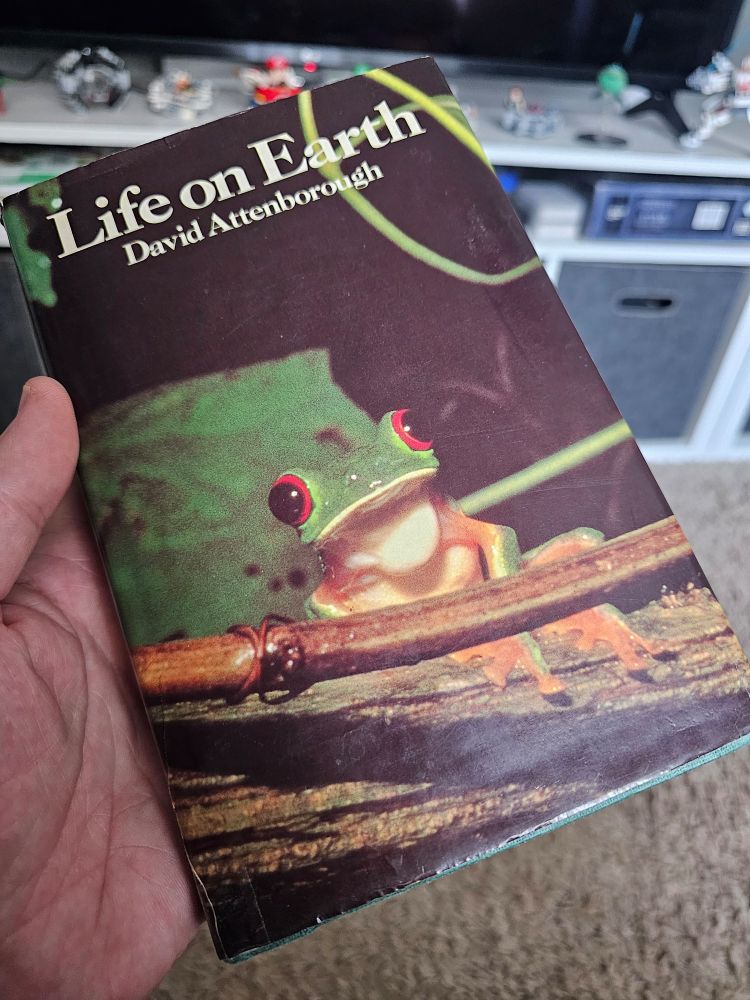 A photo showing the cover of the small-format hardback book, Life on Earth by Sir David Attenbourgh. The book cover itself shows the now-famous photo of A bright green tree frog with vibrant red and black eyes. 