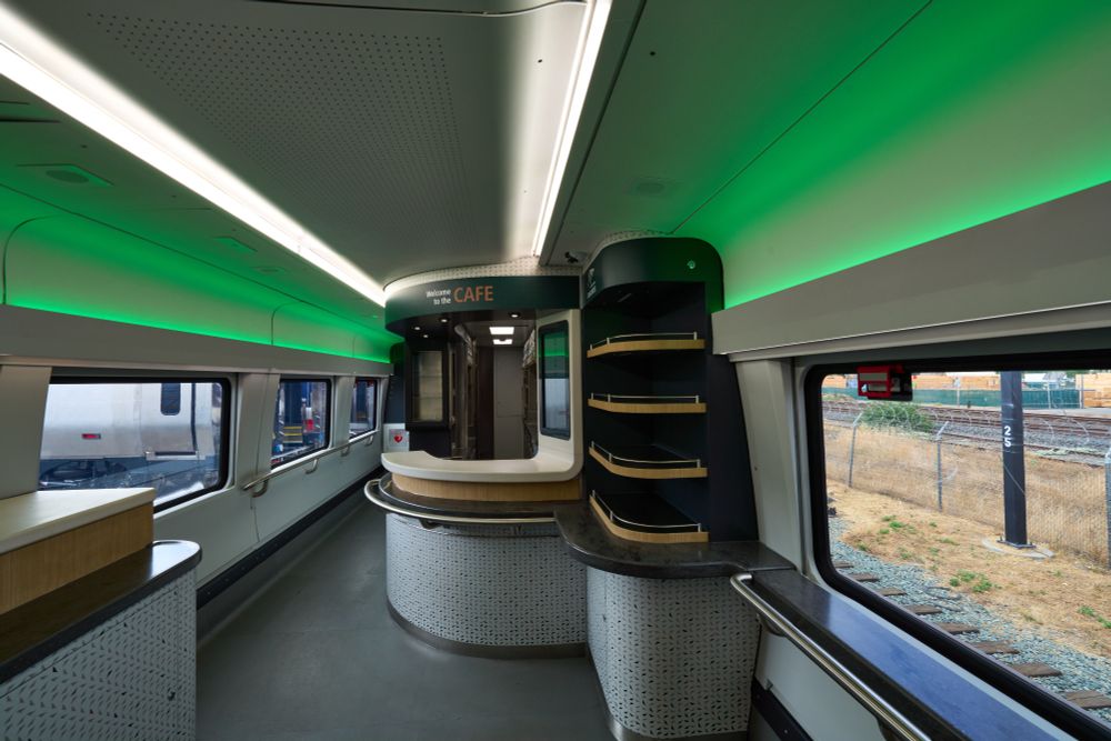 New spacious Amtrak Cascades Cafe Car features windows on both sides. A large service counter sits in the middle. Evergreen and cream colors with gold accents match the Amtrak Cascades logo, a reflection of the Pacific Northwest. White and green shadow lighting create lovely hues on the ceiling.