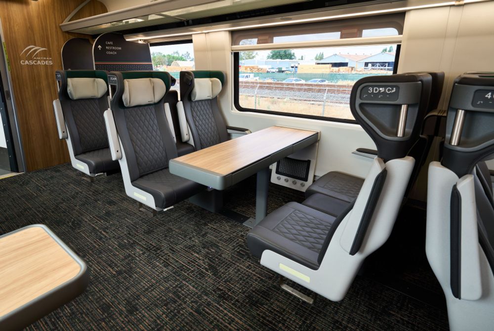 Interior photo of a new Amtrak Cascades Business Car showing two seats on one side of the car. The leather seats appear comfy with tall, puffy headrests. Some seats are facing and have tables between them. You can see the edge of a table on the single seating opposite side of the car. The windows are large to view the spectacular Northwest scenery. The floors are carpeted. The end wall has wood paneling with a white Amtrak Cascades logo.