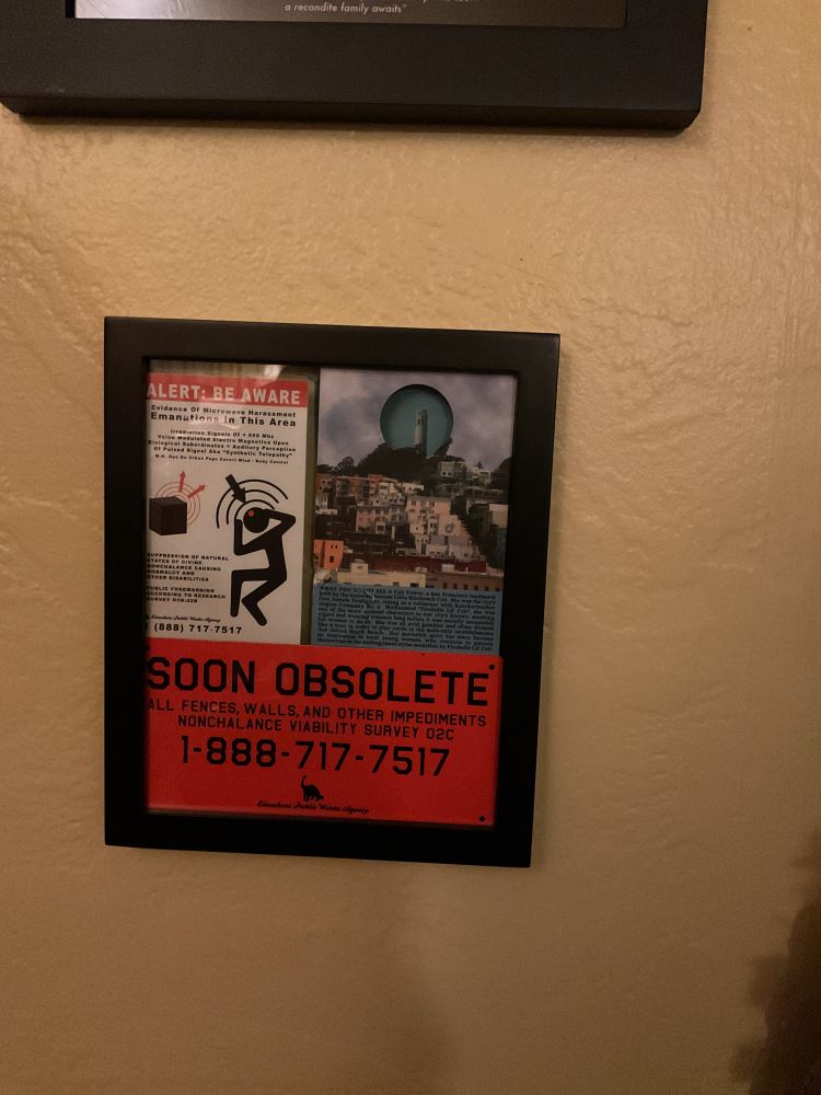 A frame holding ephemera from the Jejune Institute. A sticker warning of harassing microwave emanations, a postcard with a hole where coit tower should be(which can be seen through the hole), and a metal placard saying that fences will soon be obsolete. 