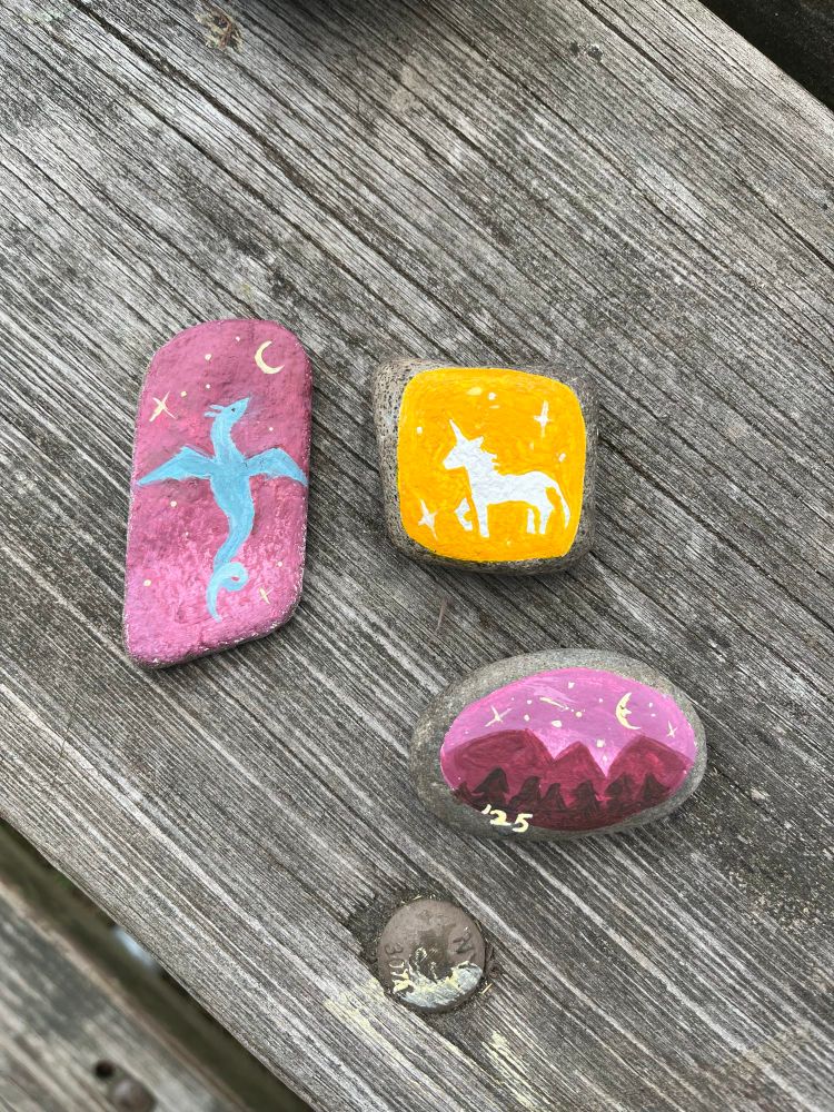 Three river stones that have been painted. The first on the left is a blue dragon against a purple night sky, the top right is a unicorn against a yellow sparkly background, and the bottom right is a mountain range with pine trees at night. In the star filled sky is a smiling moon and a shooting star. At the bottom it reads "'25"