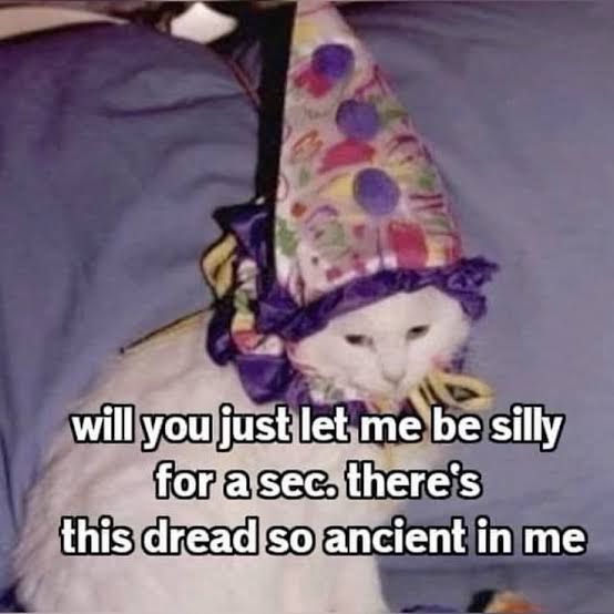 a white cat lies wearing a colorful clown hat with ruffled edges. the text over the image reads: “will you just let me be silly for a sec. there’s this dread so ancient in me”