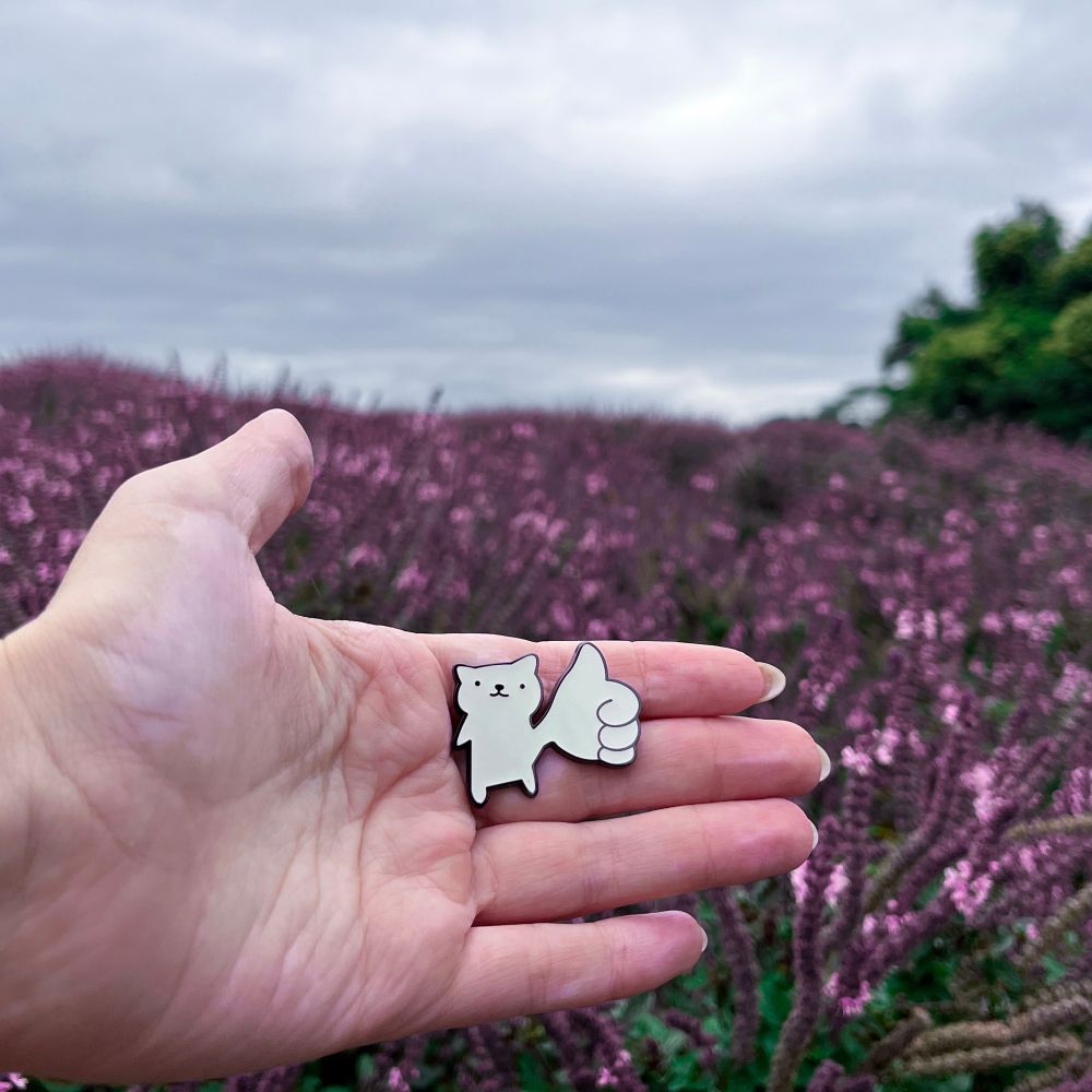 me holdind an enamel pin of a cat giving a thumbs-up gesture in front of a vibrant purple flower field