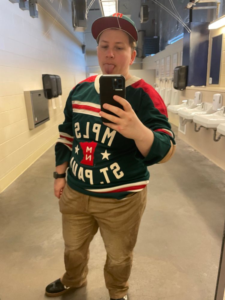 Picture of a chunky but funky white guy standing in a bathroom, sticking his tongue out and wearing a hockey jersey 