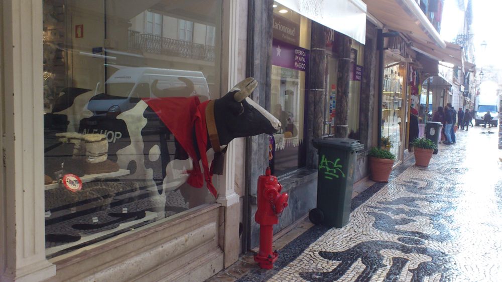 Schwarz-weiße Kuhfigur mit rotem Schal, die aus einem Schaufenster herausragt, neben einem roten Hydranten auf einem gepflasterten Bürgersteig.