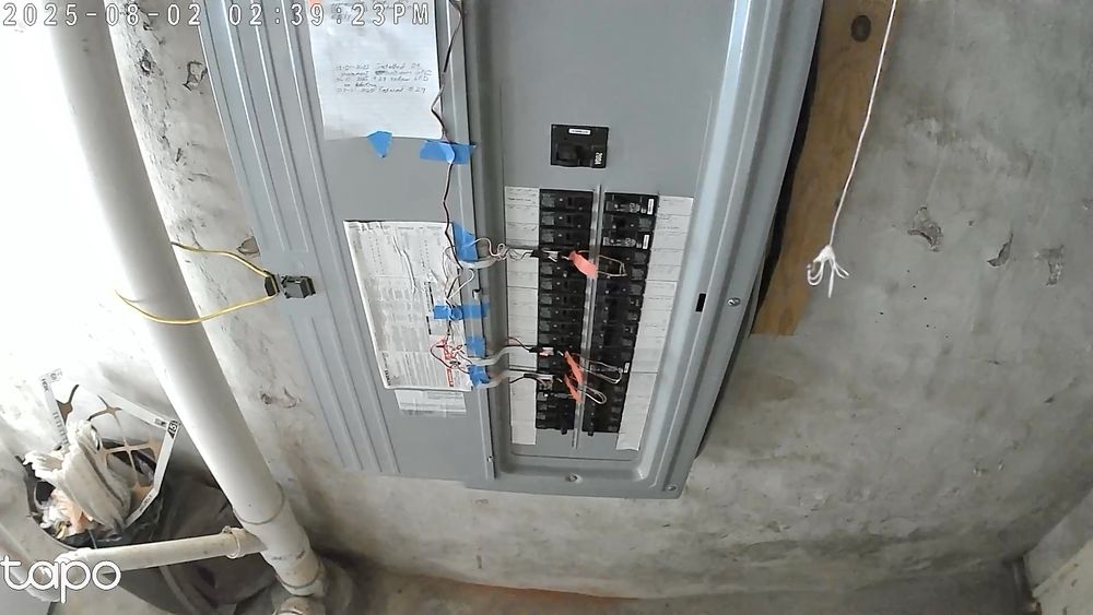A residential circuit breaker panel. Tiny switches are adjacent to 3 breakers. The switches are attached to white metal strips. The strips are held in place with 3M Command Hooks. The wires are held in place with blue masking tape. The breakers have red flags attached to the levers so the motion activated camera will detect a trip. The tiny switches make it tough to access the lever and so there is a wire loop pull handle that can be used to reset the breaker 