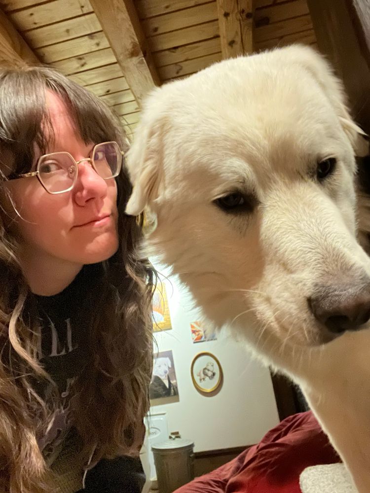 A person with long brown hair and gold rimmed glasses next to a big cute white Great Pyrenees dog