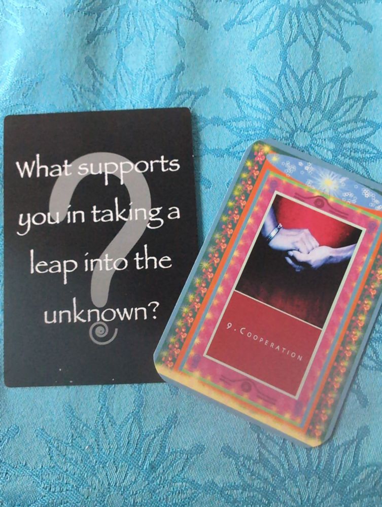 Two oracle cards on blue table cloth: On first, white text on black card with a large grey question mark reads: What supports you taking a leap into the unknown?  Second brightly decorated card shows two hands clasped on a red background with below this image the word "Cooperation" in white on a red background.