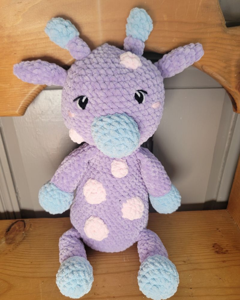 The front of a crocheted stuffie of a purple giraffe with blue hooves, snout, and antlers and pink spots.