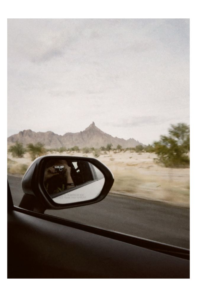 A self-portrait via the right-hand side mirror of a moving car window A desert landscape and low mountain peak are framed behind. 