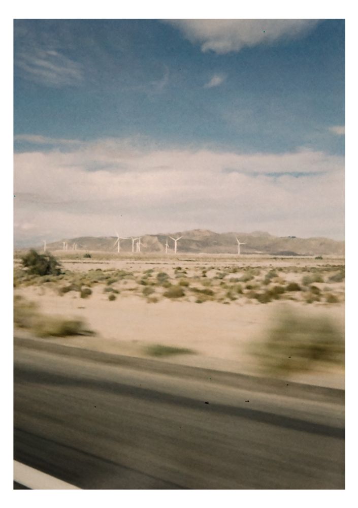 A field of wind turbines in the distance in front of a low desert mountain range, as viewed out a moving car window.
