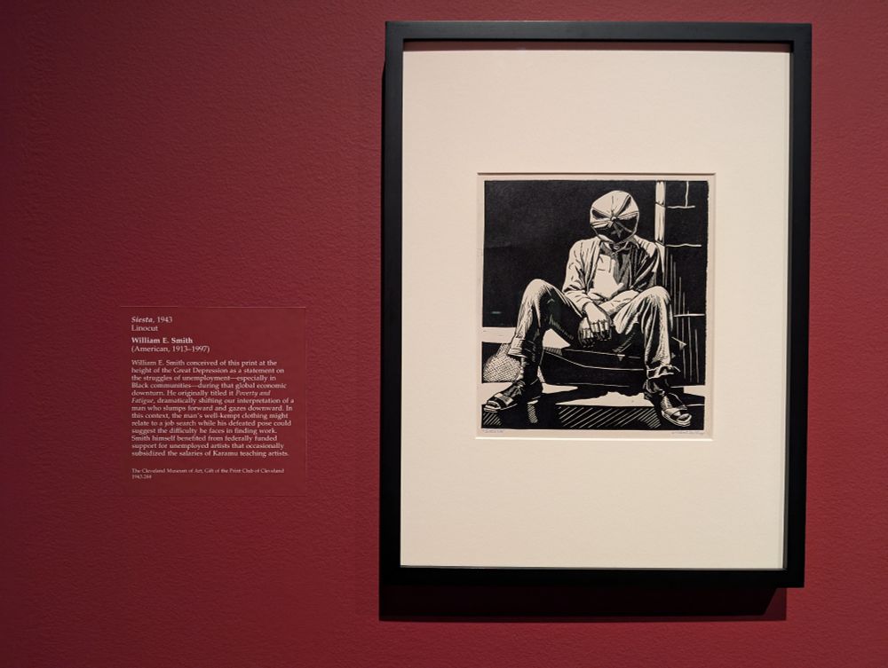 Photo of "Siesta" by American artist William E. Smith, a small 1943 monochromatic linocut print in pure black and white depicting a well-dressed man seated on some steps with his head bowed in sleep. The print is in a simple black frame on a red-painted wall. The info card is also visible to the right, and reads:

William E. Smith conceived of this print at the height of the Great Depression as a statement on the struggles of unemployment, especially in Black communities, during that global economic downturn. He originally titled it Poverty and Fatigue, dramatically shifting our interpretation of a man who slumps forward and gazes downward. In this context, the man’s well-kept clothing might relate to a job search while his defeated pose could suggest the difficulty he faces in finding work. Smith himself benefited from federally funded support for unemployed artists that occasionally subsidized the salaries of Karamu teaching artists.