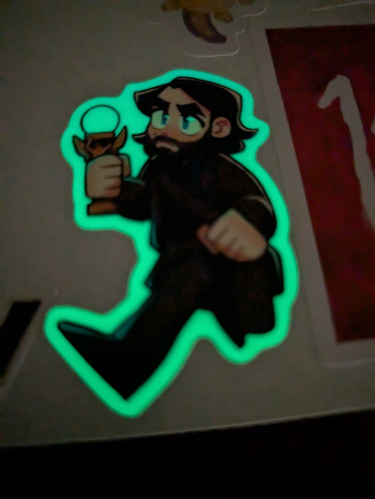 Photo taken in low light of a sticker depicting Alan Wake from the video game series of the same name. Alan Wake is a fair-skinned Caucasian man with shoulder-length dark hair dressed in a dark suit, carrying the Angel Lamp (a slim winged desk lamp topped with an exposed lightbulb) in his right hand. He is drawn in an anime-esque chibi art style and depicted mid-stride with a determined expression, and the sticker is printed on holographic, glow-in-the-dark foil that's glowing neon green in the low lighting. The highlights in his eyes are transparent, allowing the foil to show through from underneath, making his eyes look like they're shining in the dark.