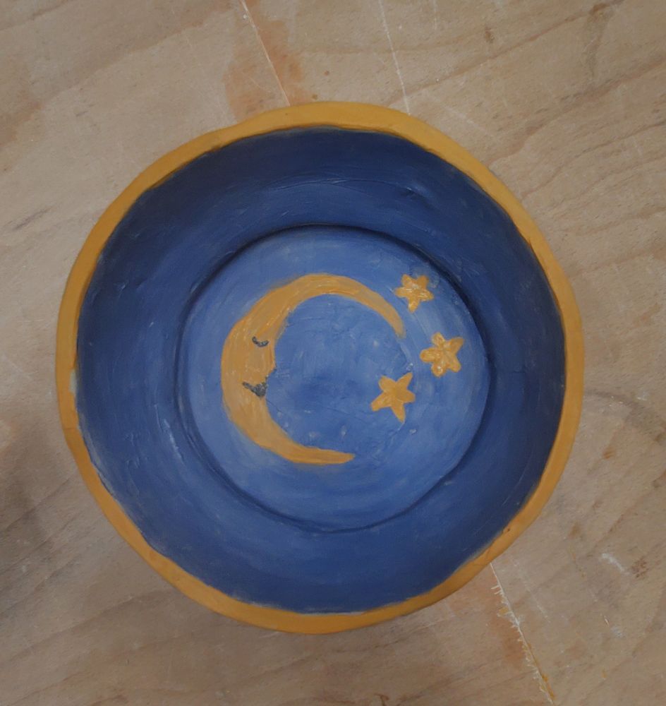photo of the bowl still on the pottery turntable on the wooden table but now finished. the inside of the bowl is dark blue with a yellow rim and on the inside bottom of the bowl is a moon with nose, lips and a closed eye and surrounded by 3 stars 